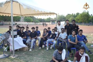 College cricket players on the field while spectators watch the Ranbhoomi 2025 match at the Railway Sports Complex.