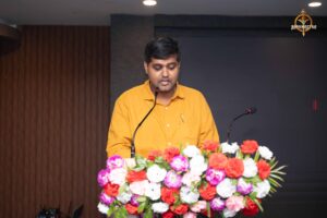 Faculty member addressing the audience with a microphone during Navya Sopna 2025 entrepreneurial event.