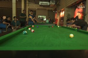 People around a pool table indoors with one taking a shot, others observing closely.