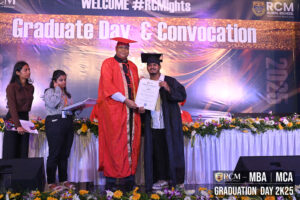 Graduate receives certificate at RCM Global Business School ceremony, stage with flowers and banner.