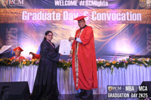 Graduate receives diploma from an official at a convocation, banner and flowers visible.