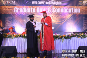 Graduate receiving certificate at ceremony with banner and floral decorations.