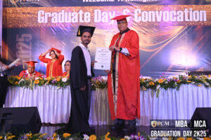 Graduate receives certificate from official in red at MBA/MCA ceremony, decorated table in background.