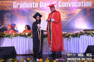 Graduate receiving certificate from official in red gown at MBA and MCA convocation event.