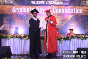 Two individuals in academic regalia at a 2025 MBA/MCA graduation ceremony, RCM event.