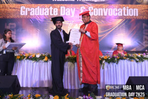 Graduate receiving certificate at RCM 2025 MBA/MCA convocation, with banners and floral decor.