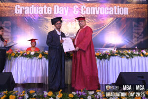 Graduate in black attire receives certificate from red-robed individual on decorated stage.
