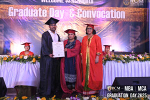 Graduate receives certificate from two women at convocation; faculty seated, floral decor.