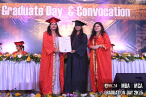 Graduate ceremony with three individuals on stage, black and red gowns, holding certificate.