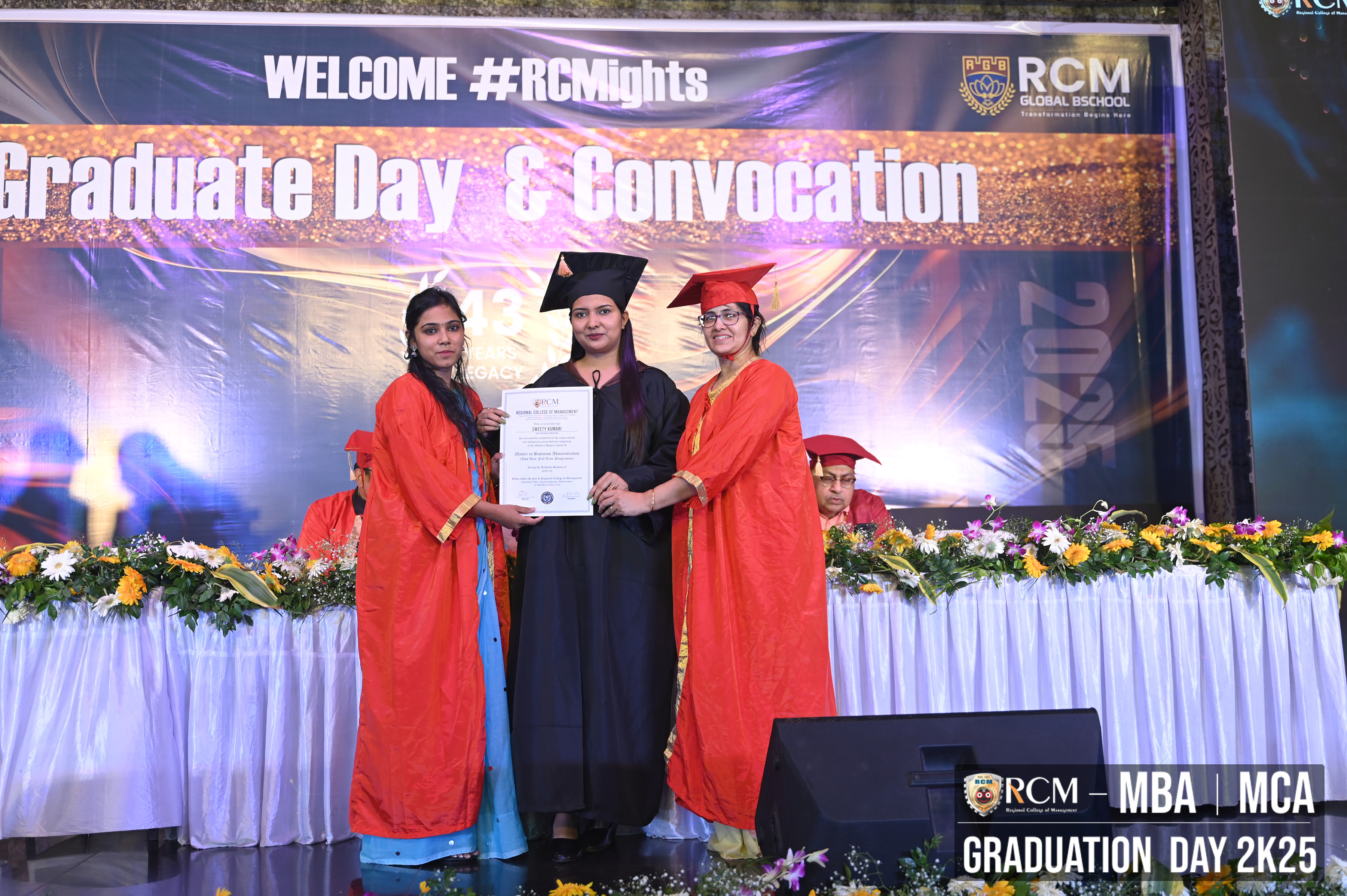Graduate receives certificate from two in red robes at RCM Global B School event with floral stage.