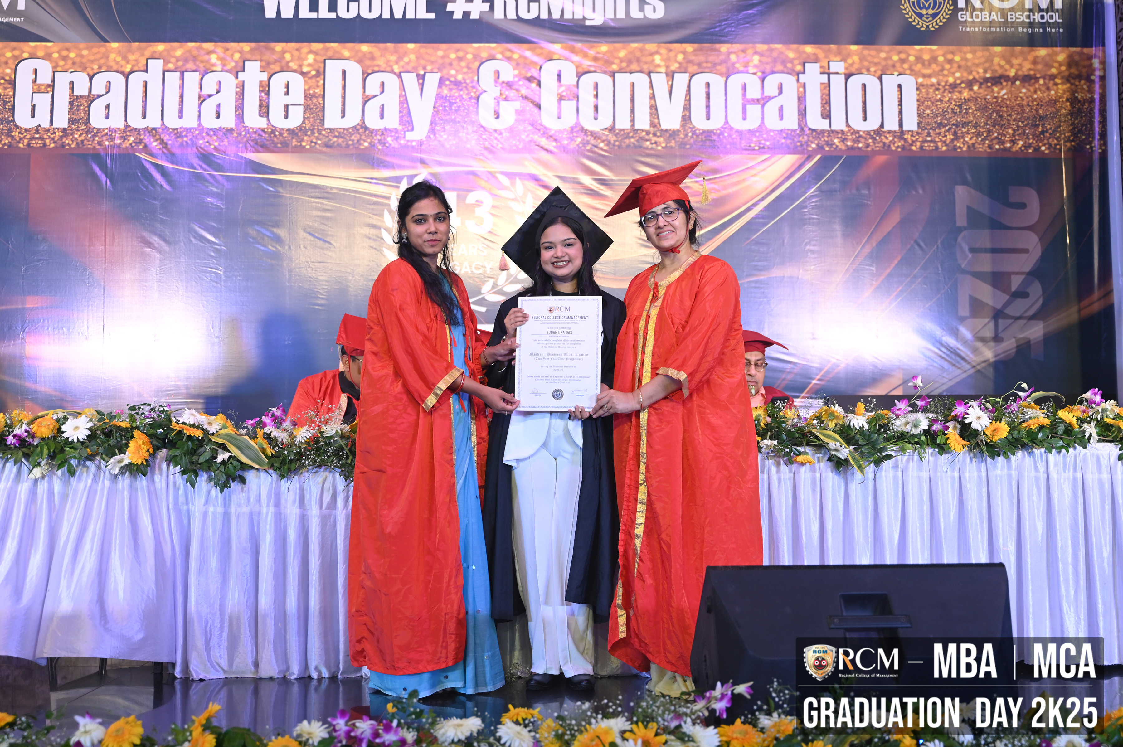 Three graduates in robes with a certificate, stage with banner "Graduate Day & Convocation."