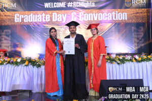 Graduate holding certificate between two people at RCM Class of 2025 ceremony stage.