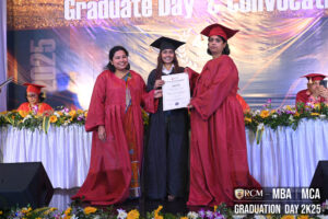 Graduation ceremony with three people in gowns, one holding a certificate, floral backdrop.