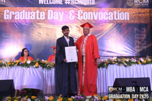 Graduate with diploma and official at RCM Global B School ceremony, stage with flowers and banners.