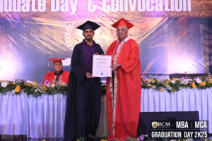 Graduate and faculty member in regalia on stage during RCM 2K25 graduation ceremony.