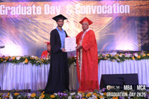 Graduate receives diploma from person in orange robe on decorated stage at convocation ceremony.