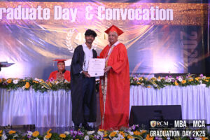 Graduate receives certificate at convocation, stage adorned with flowers, banner reads "Graduate Day & Convocation".