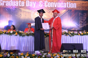 Graduate receives certificate from individual in orange robe on stage with "Graduate Day" banner.
