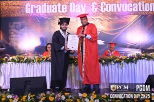 Student in gown receives certificate from faculty on stage during Graduate Day event with flowers.