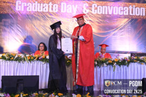 Graduate receiving certificate at convocation; decorated table, banner above, seated faculty.