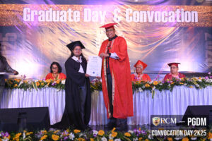Graduate receiving certificate from academic official during convocation, backdrop with flowers.
