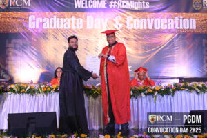 Graduate in black gown receives certificate at convocation, stage with flowers and banner.