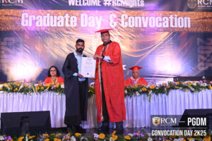 Graduate receiving certificate from official in red robe at convocation, flowers on table.