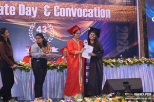 Graduation ceremony with a woman in red regalia presenting awards on a decorated stage.
