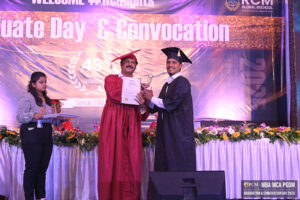 Graduate receives certificate and trophy at convocation, with decorated stage and onlooker nearby.