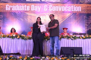 Graduation event, person in gown receiving certificate and trophy, audience in academic attire.