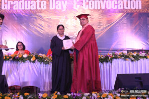 A man in a red gown awards a certificate to a woman in black at a convocation on a decorated stage.