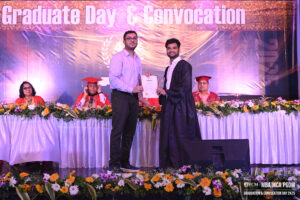 Graduate receiving certificate on stage; people in red gowns seated; backdrop reads "Graduate Day & Convocation."