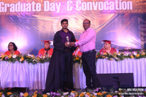 Graduate receives trophy on stage during convocation ceremony, with audience in academic attire.
