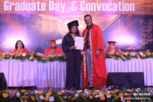Graduate with certificate at convocation event, alongside academic regalia and floral decorations.