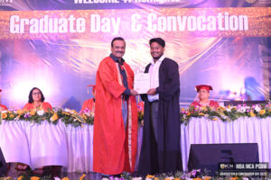 Person in black gown receives certificate from another in orange robe on decorated stage.