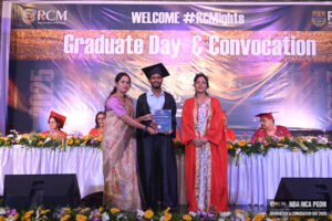 Graduate on stage in cap and gown with certificate, flanked by two women; convocation backdrop.