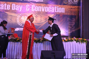 Graduation ceremony handshake between graduate in black gown and distinguished individual in red.