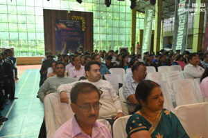 People seated in a hall with white chairs, attending a graduation event, with banners and light.