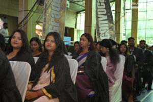 Group at an indoor graduation ceremony, wearing black gowns, with banners and large windows.
