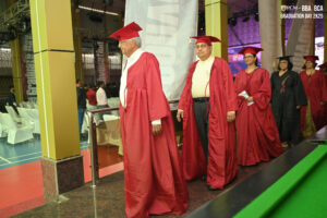 Graduates in red robes process indoors at 2025 BBA and BCA ceremony.