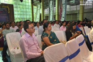 Group of people seated at an indoor graduation, dressed formally, banners in background.