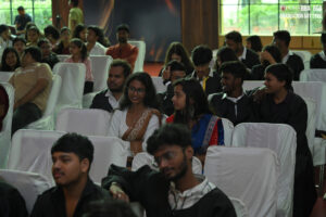 People in formal attire seated indoors on white chairs at a graduation event.