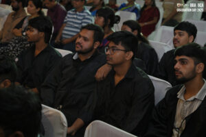 Graduates in black gowns seated attentively in an auditorium, suggesting a formal event.