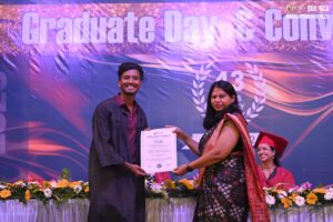 Young man in graduation gown receives certificate from woman in saree, smiling graduate seated.