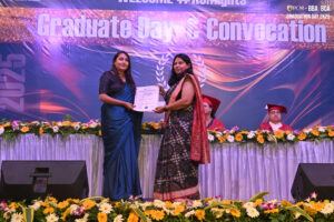 Two women on stage at graduation, one holding certificate. Floral decor and seated academics behind.