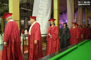 Individuals in red gowns walk at RCM BBA BCA Graduation Day 2K25, one in black robe, indoors.