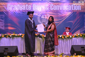 Graduate receives certificate from a woman in a sari at convocation, stage decorated with flowers.