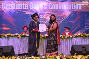 Graduation ceremony with certificate presentation, stage banner, floral decor, audience in regalia.