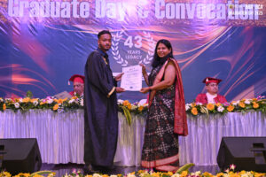 Graduate receives certificate on stage with flowers and banner, two seated in background.