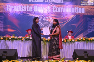 Graduation ceremony with two women exchanging a certificate on stage, floral decor, backdrop.